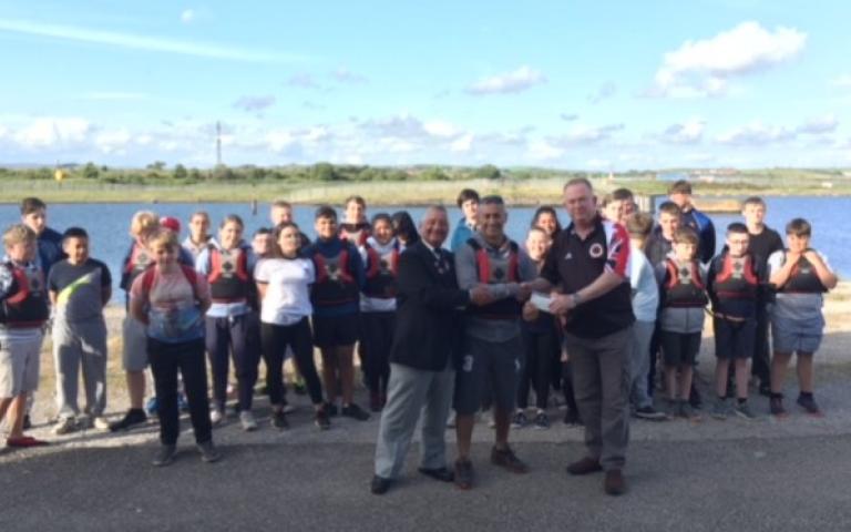 Mr Peter Fisher and Mr Ken Collins presenting a cheque to Mr Jason Zacharini, with members of the Barrow Sea Cadet Sailing Centre in the background